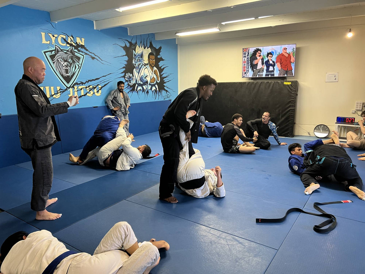 Pairs in gis roll across the blue mats at a Saturday open mat while a coach watches, with the Lycan wolf mural behind them.