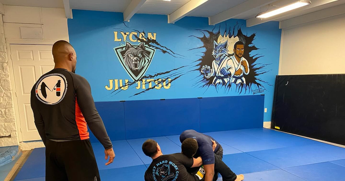 Two grapplers in rash guards work a no-gi position while a coach watches.