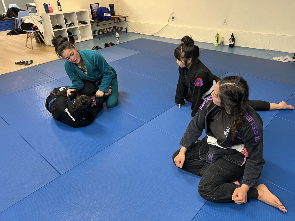 A coach in a teal gi demonstrates a guard position while two women in black gis watch on the blue mats.
