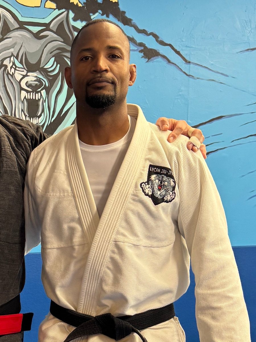 Coach Joe Monroe, in a white Lycan Jiu-Jitsu gi and black belt, in front of the Lycan wolf mural, with Coach Ken's hand on his shoulder.