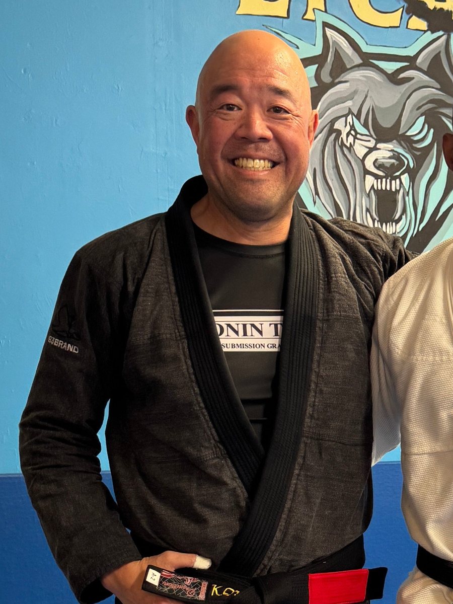 Coach Ken Kim, smiling, in a black gi and black belt in front of the Lycan wolf mural.