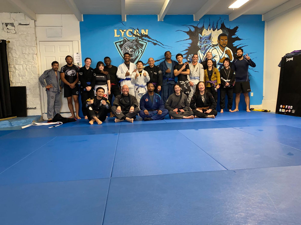 About twenty-five Lycan members, women and men of many ages and backgrounds, in gis and rash guards, posing together on the blue mats in front of the academy's wolf mural.