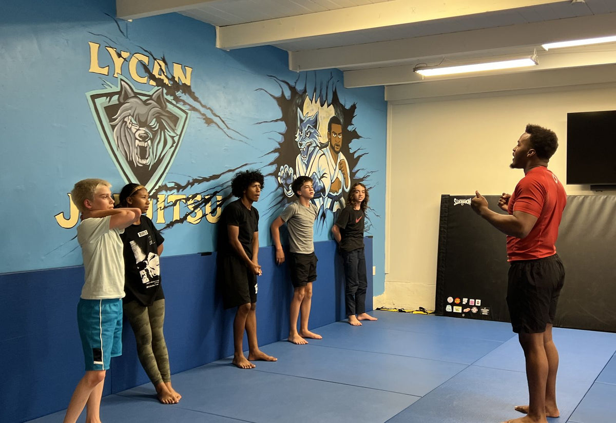 Coach Noah, in a red rash guard, talks five teen students through a drill in front of the Lycan wolf mural.